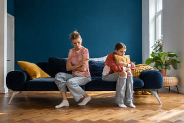 Upset teenage daughter and young woman mother sitting separately in silence on sofa at home, feeling frustrated after quarrel. Sad parent mom having misunderstandings with troubled teen girl child
