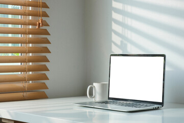 Laptop computer with empty display and cup of coffee on white table in bright office interior.