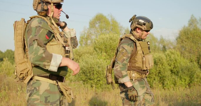 soldier taking off gloves, standing on field, his friends taking off bulletproof armor vest, jacket on hot summer day