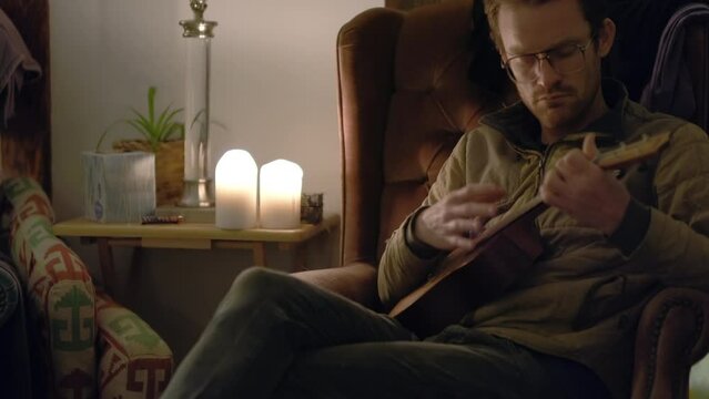 A man on an armchair at night is relaxing and playing a ukulele.