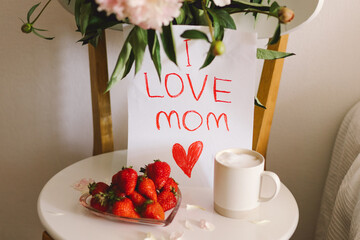 Breakfast for Mothers Day. Heart shaped white plate with fresh strawberries, cup of coffee, gift and Peonys bouquet with gift in bed. Still life composition. Happy Mother's Day.
