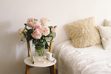Fototapeta na wymiar Still life details in home interior of living room. Open book with glasses, cup coffee and bouquet white pink peonies flowers. Read and rest. Cozy home