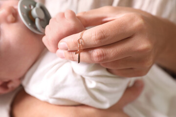 Mother holding Christian cross near newborn baby indoors, focus on hand