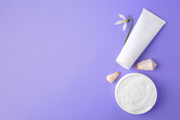 Jar and tube of cream, seashells with snowdrop flower on violet background, flat lay. Space for text