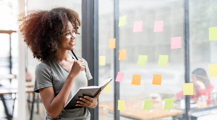 Smiling young black businesswoman holding notebook looking and thinking, standing next to a glass...