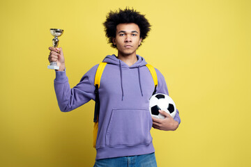 Serious proud African American man football player holding ball and trophy cup