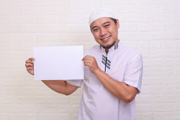 Smiling Asian muslim man in white clothes showing blank white paper for text space over white background
