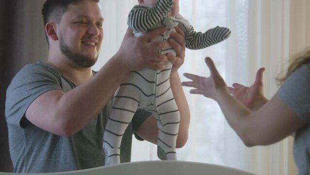 Man holds daughter or son in his arms and looks at wife. Mother puts baby in bodysuit in prepared crib. Parents plays with newborn baby arms and legs. Concept of childhood, fatherhood and family.