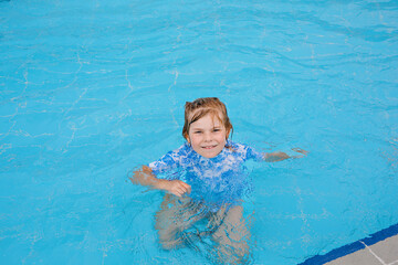 Little preschool girl playing in outdoor swimming pool in hotel resort. Child learning to swim in outdoor pool, splashing with water, laughing and having fun. Family vacations.