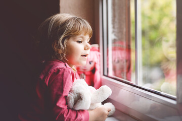 Cute toddler girl sitting by window and looking out on rainy day. Dreaming child with doll and soft...