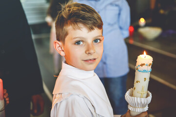 Little kid boy receiving his first holy communion. Happy child holding Christening candle. Tradition in catholic curch. Kid in a white traditional gown in a church near altar. © Irina Schmidt