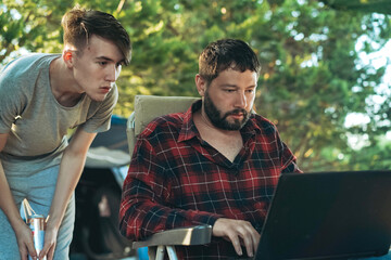 Caucasian bearded man and teenage son working wing with laptop outdoors. Camping concept