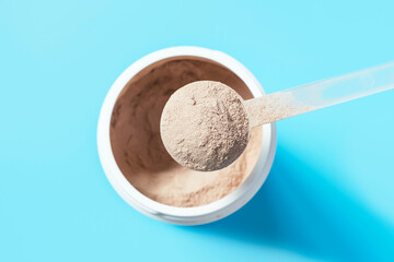 Chocolate protein powder in a scoop against a plastic jar on blue background.