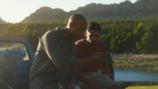 Two male friends sitting on tailgate of pick up truck on road trip in countryside drinking beer and watching sports streamed to mobile phone  - shot in slow motion