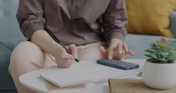 Serious young woman using smartphone touching screen and writing in notebook concentrated on business at home. Modern technology and entrepreneurs concept.