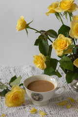 Still-life with porcelain cup of coffee, yellow garden rose flowers on pin frog in vintage vase on white lace tablecloth