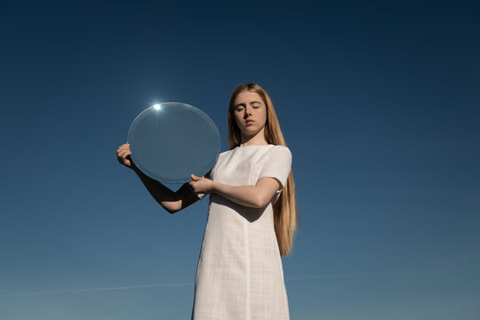 woman in white dress holding round mirror reflecting the blue sky