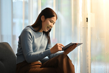 Joyful young woman sitting on sofa in cozy home, using digital tablet for online communication or shopping on internet