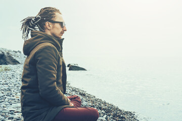 Young man with dreadlocks sits in lotus position on stone beach near sea water and meditates, enjoying peace and tranquility of nature.