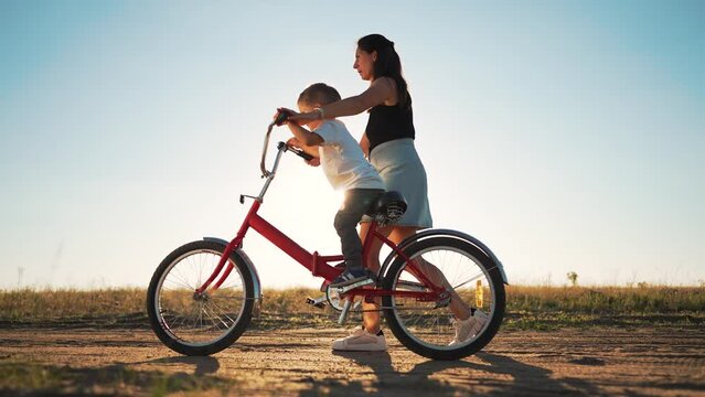 Happy family concept. Mom teaches her son to ride bike on green grass. Child rides bicycle along rural road. Green energy. Mom teaches her son to ride bike for the first time in park at sunset.