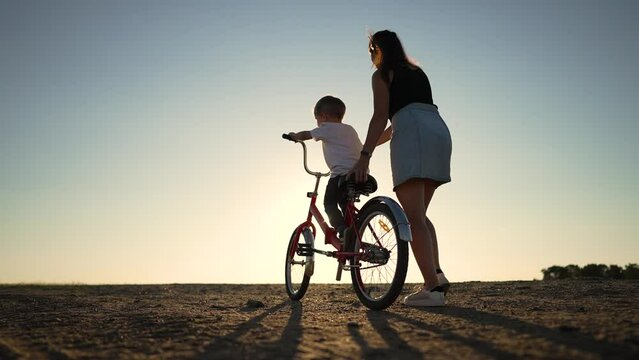 Happy family concept. Mom teaches her son to ride bike on green grass. Child rides bicycle along rural road. Green energy. Mom teaches her son to ride bike for the first time in park at sunset.