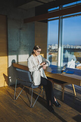 Work in a modern office. Positive young businesswoman in casual clothes and glasses sitting in the office with a mobile phone while working on a remote project using a laptop