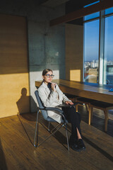 Work in a modern office. Positive young businesswoman in casual clothes and glasses sitting in the office with a mobile phone while working on a remote project using a laptop