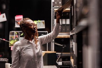 African american woman detective taking csi report file from rack with folders. Law enforcement...