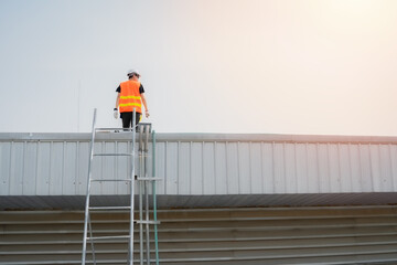 Asian engineer wearing a protective vest and a white hardhat is standing on the roof and looking to the copy space with warm light.