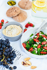 Vegan picnic food: hummus, lemonade, broccoli salad and fruits on white blanket on the grass.