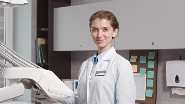 Medium portrait of young Caucasian female dentist in white lab coat smiling at camera standing by modern dental chair in modern dental office