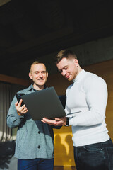 Work of two male colleagues standing in office together while exchanging ideas with laptop in meeting room and analyzing work schemes. Work in a modern, spacious office