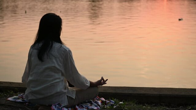 young woman practicing yoga meditation for mindfulness after work near water lake, sitting meditate on banks of canal river, meditating with closed eyes in sunset silhouette time