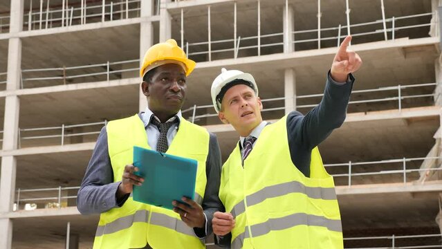 African-american and Eropean engineers standing on construction site and talking about new project. One man holding folder paper with documentation, another one pointing with finger.