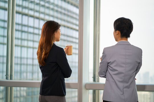 two asian businesswomen standing talking chatting by the window in office
