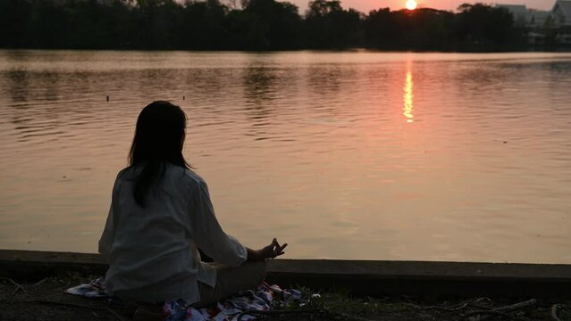 young woman practicing yoga meditation for mindfulness after work near water lake, sitting meditate on banks of canal river, meditating with closed eyes in sunset silhouette time