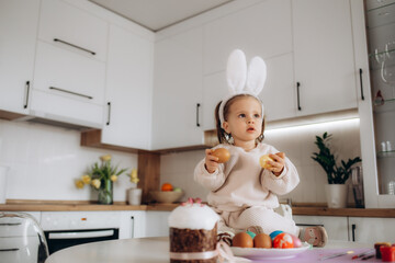 Cute little child wearing bunny ears. Girl sitting on the window with painting eggs.