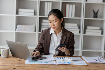 Young beautiful asian businesswoman using laptop and smart phone and calculator for doing financial mathematics on desk, tax, report, accounting, statistics and analytical research concept.