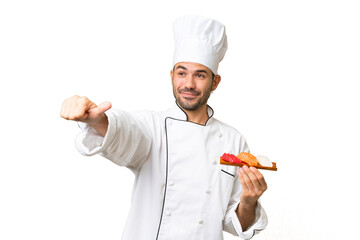 Young caucasian chef holding a sushi over isolated background giving a thumbs up gesture