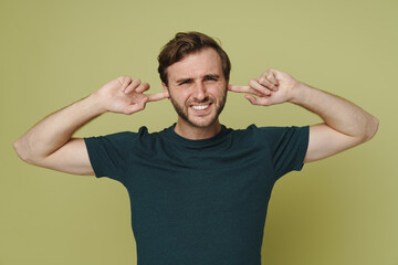 Annoyed man frowning and covering his ears isolated over green wall