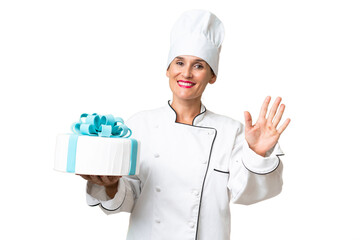 Middle-aged chef caucasian woman with a big cake over isolated background saluting with hand with happy expression
