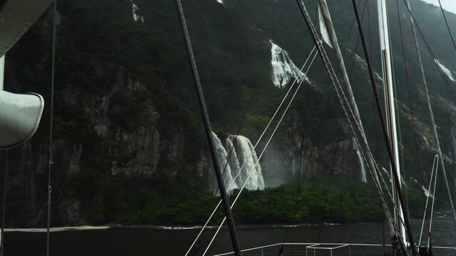 Falls cascade down steep slope of Milford Sound on rainy day; view from boat