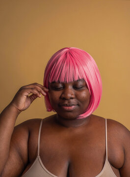 Plus size woman wearing pink bob wig posing with eyes closed against beige background