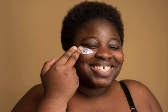 Plus size woman applying cosmetic oil on face against beige background