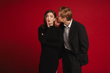 Surprised young couple gossiping isolated over red wall