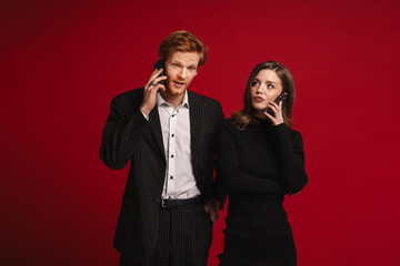 Displeased couple talking on mobile phones while standing isolated over red background