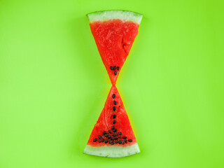 Triangle slices of delicious red watermelon in shape of hourglass clock on a green background, creative idea. Summer time concept. Top view flat lay