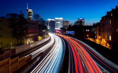 Fototapeta na wymiar Curved Motorway A40 called “Ruhrschnellweg“ at blue hour after sunset in Essen City Ruhr Basin Germany with six lanes, bright light traces, sky gradient and skyline silhouettes of office towers.