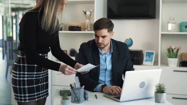 Angry young man, manager scolding his employee in the office for a poorly done job, gesturing emotionally, sitting on a laptop, throwing papers at a confused pretty woman in business clothes.