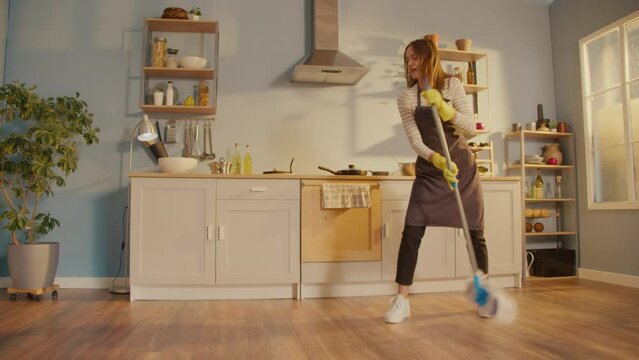 Creative Woman is Washing Floor With Mop Singing and Dancing in Kitchen at Home. Happy Young Woman Cute Housewife is Listening to Music and Dancing in Front of the Fan. Household Chores Concept.